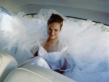 Braut sitzt im Tüllkleid auf der Rückbank im Auto und lächelt | © Getty Images/Laurence Monneret