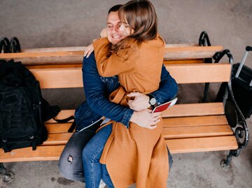 Frau sitzt am Bahnhof bei ihrem Partner auf dem Schoß und umarmt ihn | © Getty Images/Nikola Stojadinovic