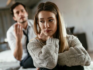 Frau sitzt nachdenklich auf dem Bett, während ihr Partner auf sie einredet | © Getty Images/Boris Jovanovic