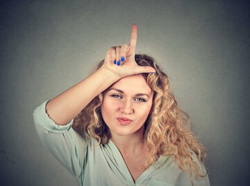 Blonde Frau zeigt mit den Fingern den Buchstaben L auf ihrer Stirn | © Getty Images/SIphotography