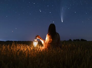 Frau sitzt nachts mit einer Laterne auf einer Wiese und beobachtet Sternschnuppe | © Getty Images/Anton Petrus