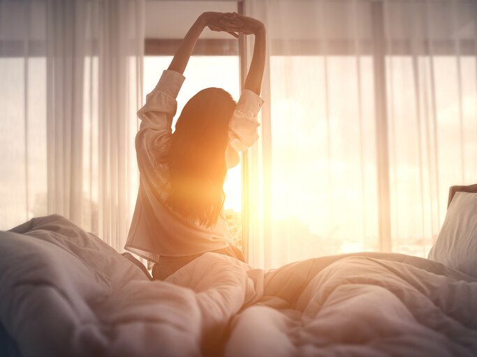 Eine Frau streckt sich im Bett bei Sonnenaufgang | © GettyImages/oatawa