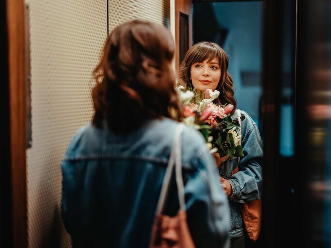 Frau in einem verspiegelten Aufzug mit einem Blumenstrauß in der Hand | © Getty Images/miniseries