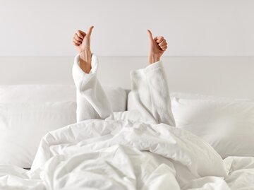 Frau in Bademantel liegt unter der Bettdecke und zeigt mit Daumen nach oben | © AdobeStock/Syda Productions