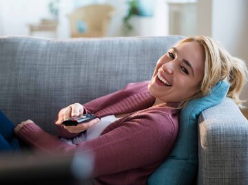 Junge Frau liegt auf einem Sofa mit der Fernbedienung in der Hand | © Getty Images/JGI/Jamie Grill