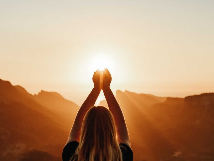 Junge Frau in spiritueller Pose, die das Licht vor den Bergen hält | © Getty Images/DianaHirsch