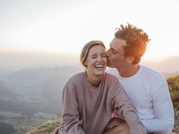 Verliebtes junges Paar sitzt vor einem Sonnenuntergang. Mann küsst Frau auf die Wange und sie lächelt | © Getty Images/Westend61