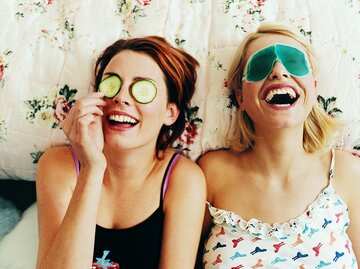 Zwei junge Frauen lachen auf dem Bett zusammen. Eine Frau hat Gurkenscheiben auf ihren Augen, während die andere Frau Augenpads trägt | © Getty Images/Digital Vision.