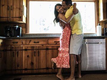 Verliebtes Paar umarmt sich in der Küche. Mann küsst Frau auf die Wange, sie lächelt. | © Getty Images/MoMo Productions
