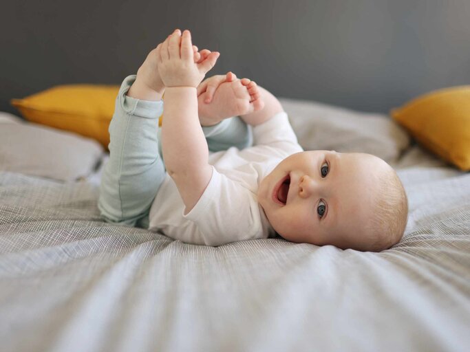 Süßes Baby liegt auf dem Bett und greift an seine Füße | © Getty Images/Catherine Delahaye