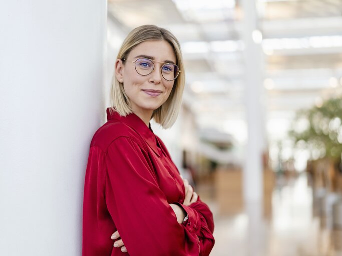 Junge blonde Frau trägt Brille, rote Bluse und lächelt in die Kamera. Ihre Arme sind verschränkt und sie lehnt sich gegen eine Wand | © Getty Images/Westend61