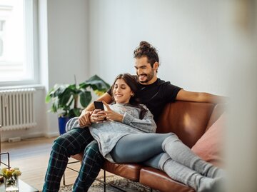 Junger Mann sitzt auf der Couch, seine Freundin hat ihren Kopf auf seinem Schoß und beide schauen sich lächelnd etwas auf dem Handy an | © Getty Images/Luis Alvarez