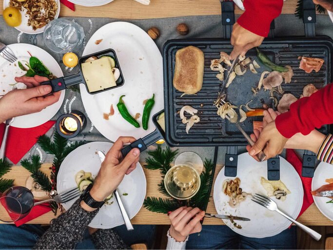 Familie bereitet verschiedene Gerichte auf Raclette-Grill auf Holztisch | © Getty Images/golero