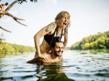 Mann und Frau lachen zusammen im See, während sie auf seine Schultern klettert | © Getty Images/Luis Alvarez