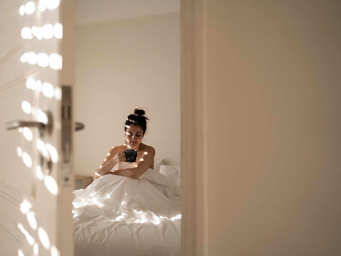 Frau sitzt im Bett und trinkt einen Kaffee bei geöffneter Tür | © Getty Images/Natalia Romero/EyeEm