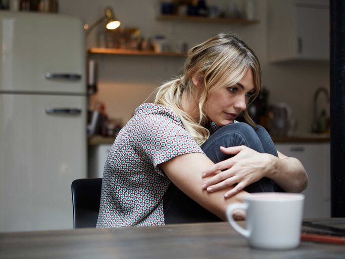 Blonde Frau sitzt mit angezogenen Beinen am Tisch und ist traurig. | © Getty Images / Westend61