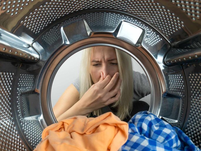 Junge blonde Frau schaut auf die Wäsche in der Waschmaschine und hält ihre Nase gegen einen unangenehmen Geruch | © Getty Images/Olga Ihnatsyeva