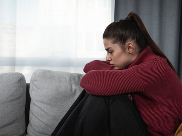 Frau sitzt nachdenklich auf einer Couch und hat die Knie an ihren Oberkörper gezogen | © Getty Images/Srdjanns74
