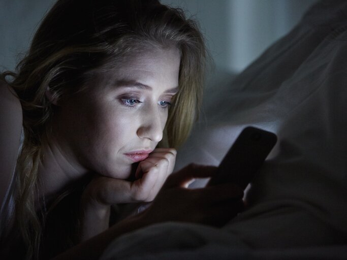 Frau liegt im Dunkeln auf dem Bett und schaut auf ihr Handy  | © Getty Images/Lilly Bloom