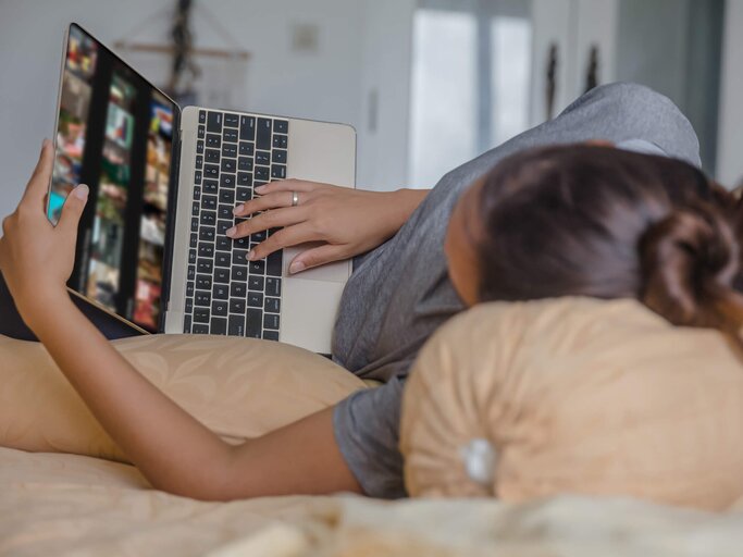 Frau liegt seitlich im Bett und guckt Serien am Laptop | © gettyimages.de / Kanawa_Studio