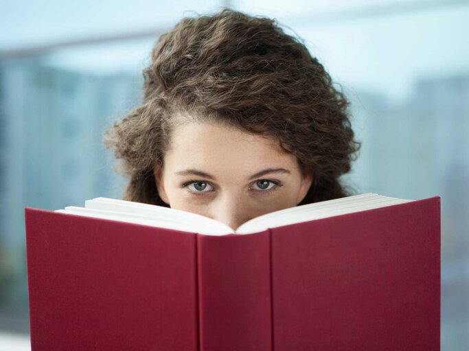 Frau blickt hinter einem Buch hervor | © Getty Images/Westend61