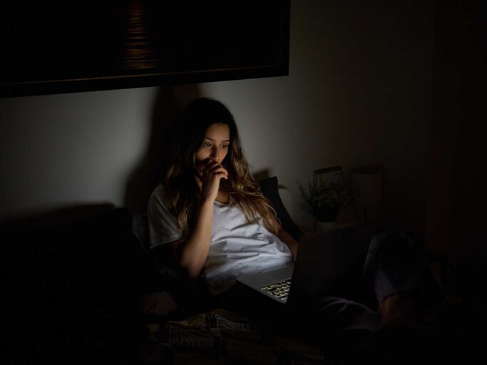 Frau schaut einen Film auf ihrem Laptop und sitzt im Dunkeln auf dem Bett | © Getty Images/Javier Sanz