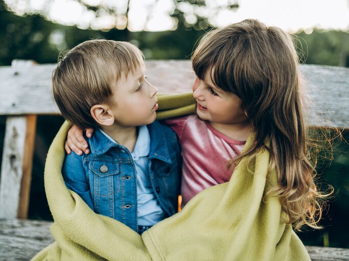 Ein kleines Mädchen und ein kleiner Junge sitzen nebeneinander und umarmen sich | © GettyImages/Liliya Krueger