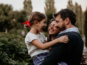 Ein Vater hält seine junge Tochter auf dem Arm, die Mutter steht daneben und alle freuen sich miteinander. | © Getty Images / Studio4