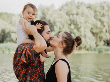 Junge Familie steht am Wasser, Vater und Mutter küssen sich. | © Getty Images / Senserini Lucrezia