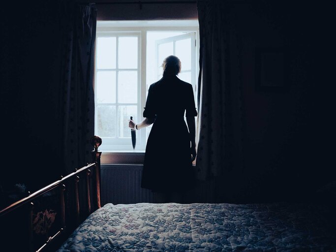 Frau steht mit einem großen Messer am Fenster und wartet. | © Getty Images / Mimi Van Praagh