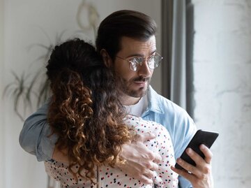 Mann umarmt seine Frau und schaut hinter ihrem Rücken auf sein Handy. | © Getty Images / fizkes