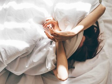 Frau liegt mit dem Kopf unter der Decke im Bett und hält sich mit den Armen den Kopf. | © Getty Images / Maria Korneeva