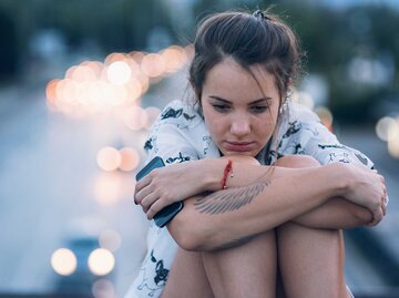 Frau sitzt mit angezogenen Beinen auf der Straße und hat ist traurig. | © Getty Images / Mixmike