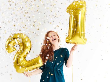 Frau steht im Konfetti-Regen mit zwei Zahlenluftballons. | © Getty Images / Westend61