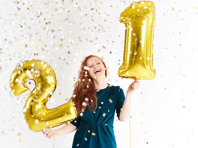 Frau steht im Konfetti-Regen mit zwei Zahlenluftballons. | © Getty Images / Westend61