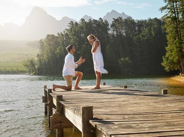 Junger Mann macht junger Frau auf einem Steg am See einen Heiratsantrag. | © Getty Images / PeopleImages