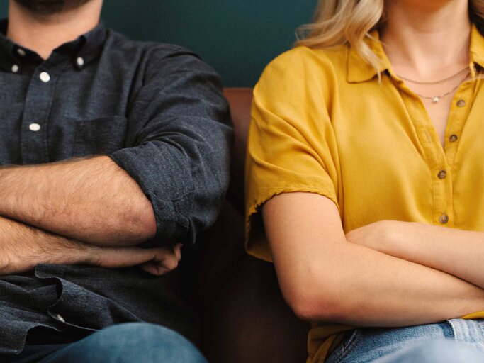 Mann und Frau sitzen mit verschränkten Armen nebeneinander auf dem Sofa | © Getty Images/shapecharge