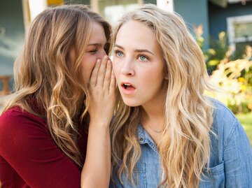 Eine junge Frau flüstert ihrer Freundin etwas ins Ohr, diese schaut schockiert. | © Getty Images / Jessica Peterson