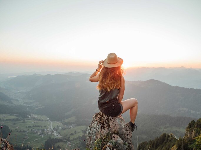 Frau mit Hut sitzt auf einem Gipfel und schaut in den Sonnenuntergang | © Getty Images/Westend61