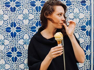 Junge Frau steht vor einem Mosaik-Hintergrund und isst ein Eis in der Waffel. | © Getty Images / Cavan Images