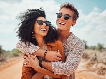 Glückliches Paar umarmt sich lachend in den Dünen einen Strandes. | © Getty Images / PeopleImages