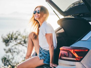 Junge Frau sitzt lachend im Kofferraum ihres Autos | © Getty Images/Maria Korneeva