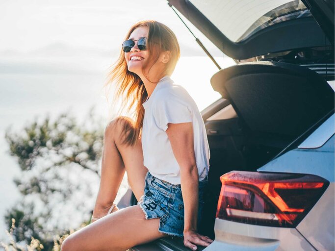 Junge Frau sitzt lachend im Kofferraum ihres Autos | © Getty Images/Maria Korneeva