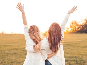 Zwei Frauen stehen Arm in Arm auf einem Feld im Sonnenuntergang und strecken ihre Arme in die Luft. | © Adobe Stock/Dima Aslanian