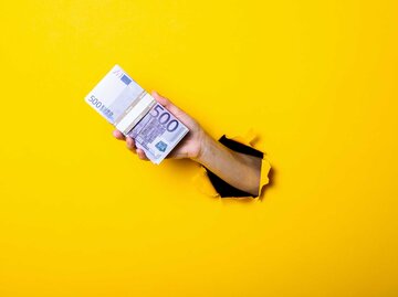 Eine Hand kommt aus einer gelben Wand und hält ein Bündel mit 500 Euro Scheinen. | © Adobe Stock/Alex