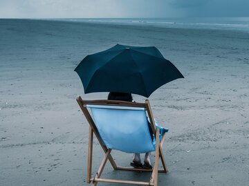 Frau sitzt in Liegestuhl mit Regenschirm am Strand | © Getty Images/Dan Brownsword