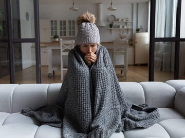 Frau sitzt in Wolldecke und mit Mütze frierend auf Couch | © Getty Images/fizkes