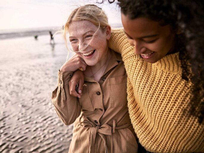 Zwei junge Frauen laufen Arm in Arm am Strand entlang.  | © Getty Images / Oliver Rossi