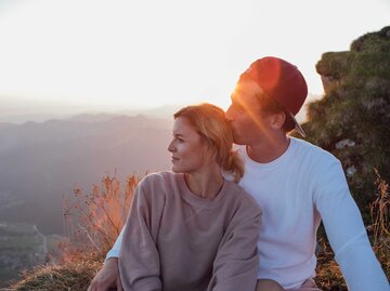 Junges, glückliches Paar sitzt beim Sonnenuntergang zusammen, der Mann gibt der Frau einen Kuss auf den Hinterkopf. | © Getty Images / Westend61