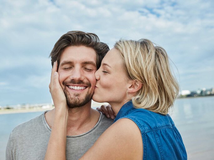 Frau mit blonden Haaren gibt einem Mann mit braunen Haaren einen leidenschaftlichen auf die Backe. | © Getty Images / Westend61